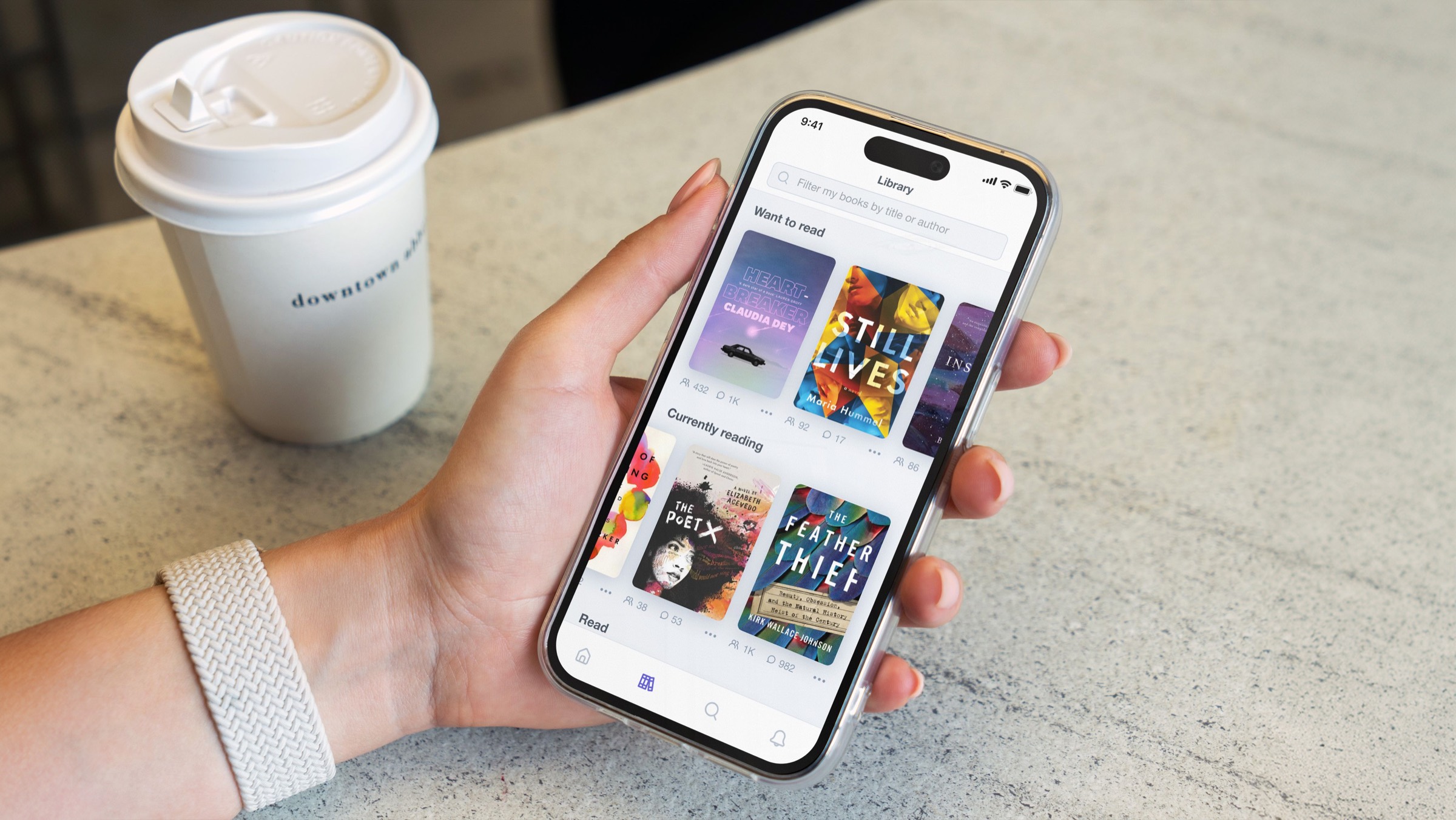 A hand holding a phone that shows a Byzans library of book covers, next to a coffee cup